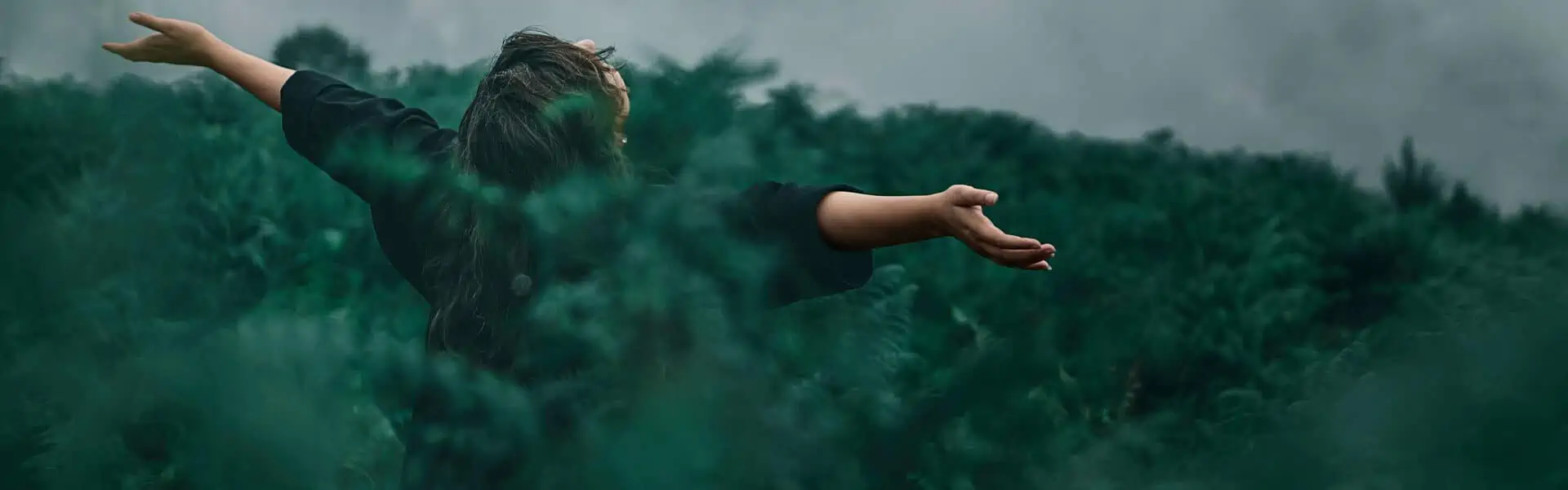 A woman stands with her arms outstretched and her face turned towards the sky in a sea of green ferns.